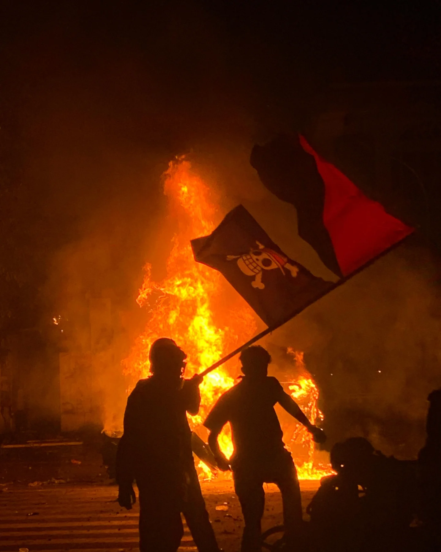The anarchist black and red bisected flag flying with the One Piece pirate flag while a fire rages in the background